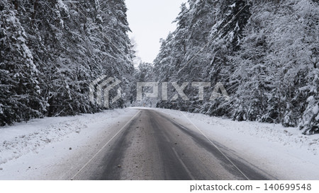 Icy two-lane road stretching into distance through dense evergreen forest heavily coated with fresh snow, compacted snow shoulders and slushy tire tracks on 140699548