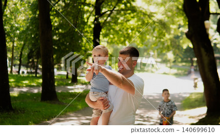 Young father holding toddler boy at shoulder height while pointing ahead along sunlit tree-lined park path; dappled afternoon light filters through leafy Young father holding toddler boy at shoulder height while pointing ahead along sunlit tree-lined park path; dappled afternoon light filters through leafy 140699610