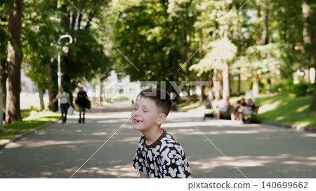 Young boy wearing a black and white patterned shirt smiling and leaning forward on a paved park pathway surrounded by tall leafy trees, dappled sunlight Young boy wearing a black and white patterned shirt smiling and leaning forward on a paved park pathway surrounded by tall leafy trees, dappled sunlight 140699662