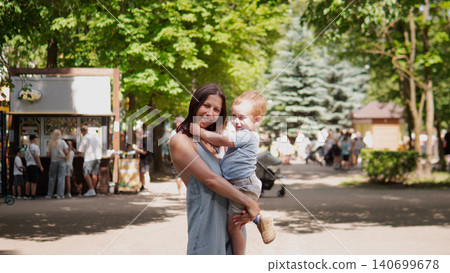 Smiling mother holds her toddler in arms while standing on a sunlit tree-lined park path with blurred market stalls and people in the background, warm natural Smiling mother holds her toddler in arms while standing on a sunlit tree-lined park path with blurred market stalls and people in the background, warm natural 140699678