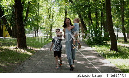 Woman wearing a light summer dress walks down a tree-lined paved path in a green park, holding a toddler in one arm while an older child walks beside her 140699684