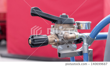 Close view of a metal industrial valve assembly with black handle and attached blue hoses against a red background, showing stainless fittings, clamps, and mechanical detail in a 140699687