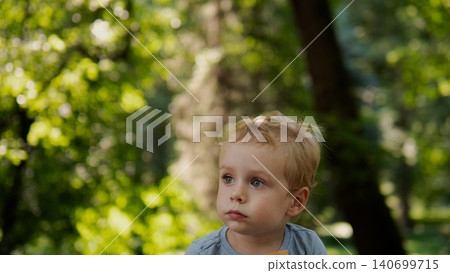 Young blond toddler seated outdoors amid sunlit green foliage, wearing a light blue shirt and looking slightly to the side with a pensive expression; soft Young blond toddler seated outdoors amid sunlit green foliage, wearing a light blue shirt and looking slightly to the side with a pensive expression; soft 140699715