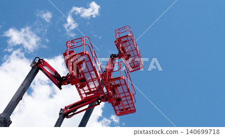 Multiple red aerial work platforms are raised high against a bright blue sky with scattered white clouds, creating a strong industrial construction scene with open space 140699718