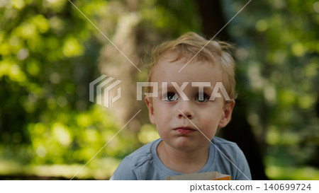 Close-up of a toddler with light hair and blue eyes looking upward while holding a small cardboard box, framed against a softly blurred green park background Close-up of a toddler with light hair and blue eyes looking upward while holding a small cardboard box, framed against a softly blurred green park background 140699724