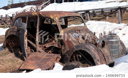 Severely rusted four-door sedan stripped of parts, doors ajar and interior exposed, surrounded by patchy melting snow and dead grass Severely rusted four-door sedan stripped of parts, doors ajar and interior exposed, surrounded by patchy melting snow and dead grass 140699733