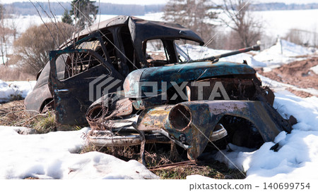 Heavy rusted vintage car wreck sits abandoned in melting snow within rural landscape, torn roof and crushed front end revealing corroded metal panels, broken Heavy rusted vintage car wreck sits abandoned in melting snow within rural landscape, torn roof and crushed front end revealing corroded metal panels, broken 140699754