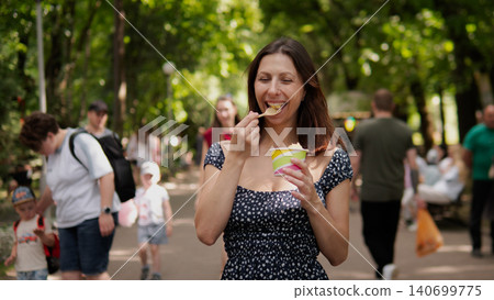Smiling woman in a patterned summer dress eating ice cream from a paper cup while standing on a tree-lined pedestrian path 140699775