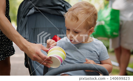 Young toddler seated in a stroller reaches toward a brightly colored stacking cup offered by an adult hand Young toddler seated in a stroller reaches toward a brightly colored stacking cup offered by an adult hand 140699823