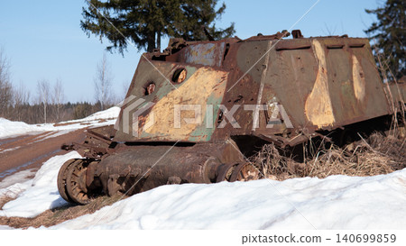 Large rusting armored vehicle hull half-buried in melting snow beside dirt road, surrounded by dry grass and pine trees under clear blue sky; metal surfaces 140699859
