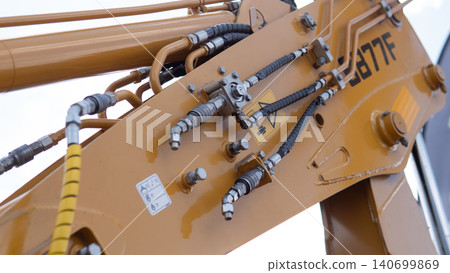 Detailed closeup of a yellow excavator arm with hydraulic hoses, metal fittings, bolts, and mechanical joints against a bright sky, showing industrial construction equipment 140699869
