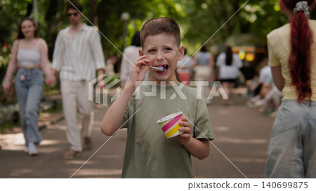 Young boy holding a colorful cup of ice cream and spoon while eating on a tree-lined pedestrian path, surrounded by casually dressed people walking in the Young boy holding a colorful cup of ice cream and spoon while eating on a tree-lined pedestrian path, surrounded by casually dressed people walking in the 140699875