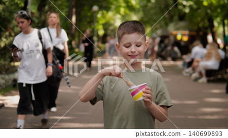 Young boy stands on a tree-lined park path enjoying a cup of ice cream with a wooden spoon, sunlight filtering through leaves and pedestrians walking 140699893