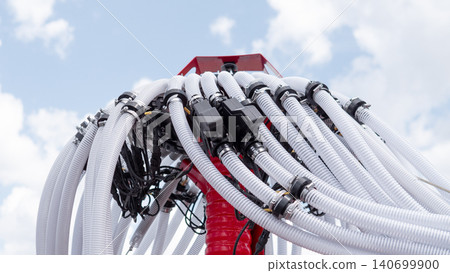 Close-up view of a red industrial machine topped with many white flexible hoses, black connectors, and bundled cables rising into a bright blue sky with soft clouds, creating a 140699900