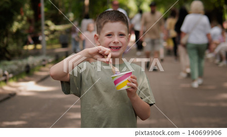 Young boy standing on a shaded tree-lined park walkway, smiling while holding a colorful paper cup of ice cream and scooping with a wooden spoon Young boy standing on a shaded tree-lined park walkway, smiling while holding a colorful paper cup of ice cream and scooping with a wooden spoon 140699906