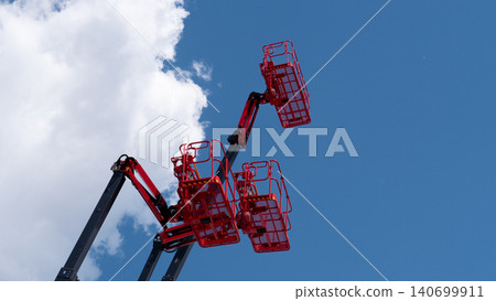 Two red boom lifts with raised baskets reach into a clear blue sky beside a large white cloud, creating an industrial construction scene with open space and bright daylight Two red boom lifts with raised baskets reach into a clear blue sky beside a large white cloud, creating an industrial construction scene with open space and bright daylight 140699911