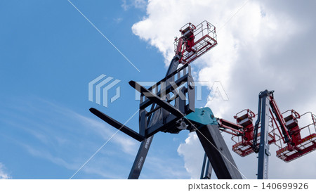 Large aerial work platforms with extended booms and red safety baskets rise overhead in an industrial setting beneath a bright blue sky and white clouds, showing heavy equipment Large aerial work platforms with extended booms and red safety baskets rise overhead in an industrial setting beneath a bright blue sky and white clouds, showing heavy equipment 140699926