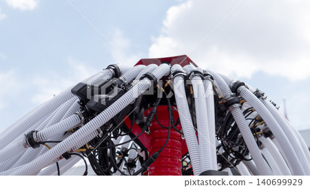 Close view of a complex industrial machine with many flexible white hoses, black cables, and a red central structure set outdoors against a cloudy sky. The equipment suggests 140699929