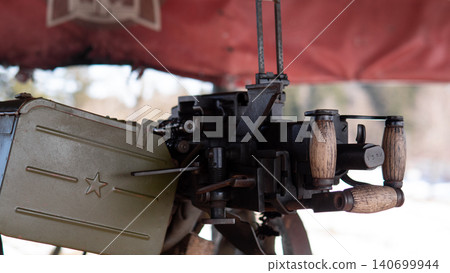 Close view of vintage heavy machine gun mounted on tripod beneath red canopy, metal ammunition box attached to side, wooden dual grips and mechanical feed 140699944