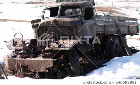 Weathered midcentury flatbed truck with corroded metal cab and wooden stake bed sitting partially collapsed in melting snow on rural field; broken windshield 140699981