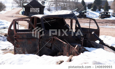 Burned rusted car shell with open front doors sits in melting snow at rural scrapyard, showing exposed chassis, missing windows, charred metal, and quiet Burned rusted car shell with open front doors sits in melting snow at rural scrapyard, showing exposed chassis, missing windows, charred metal, and quiet 140699993