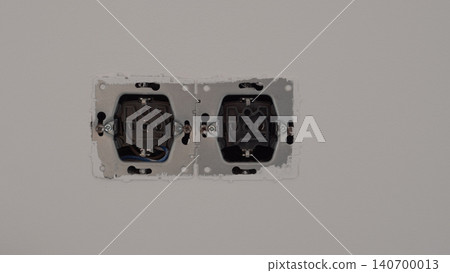 Two unfinished electrical outlet openings are mounted side by side on a plain gray wall, showing exposed inner housings and metal mounting plates in a simple interior setting Two unfinished electrical outlet openings are mounted side by side on a plain gray wall, showing exposed inner housings and metal mounting plates in a simple interior setting 140700013