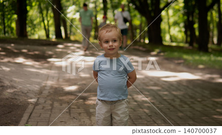 Small boy stands centered on a paved tree-lined park walkway, hands behind back and a gentle smile 140700037