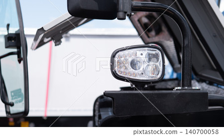Close-up view of a vehicle side mirror and bright LED work light mounted on a dark vehicle body, with a blurred white background that suggests an industrial or transport setting Close-up view of a vehicle side mirror and bright LED work light mounted on a dark vehicle body, with a blurred white background that suggests an industrial or transport setting 140700058