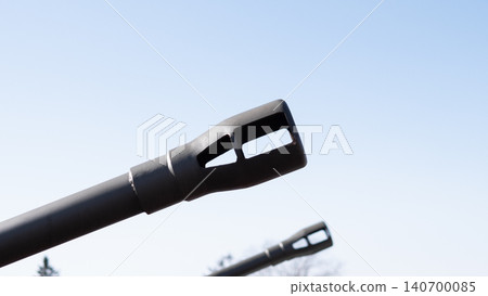 Low-angle closeup shows matte black artillery barrel muzzle pointing upward against pale blue sky, with blurred secondary barrel and faint treeline in Low-angle closeup shows matte black artillery barrel muzzle pointing upward against pale blue sky, with blurred secondary barrel and faint treeline in 140700085