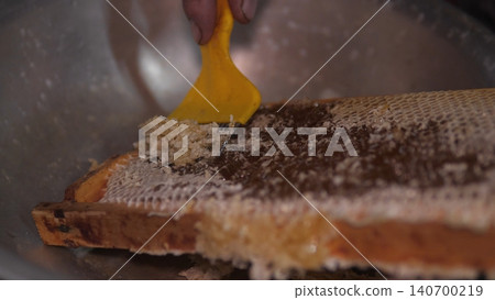 A close-up view of a hand using a yellow scraper to remove honeycomb and wax from a frame into a metal bowl, with textured comb, sticky residue, and warm natural tones A close-up view of a hand using a yellow scraper to remove honeycomb and wax from a frame into a metal bowl, with textured comb, sticky residue, and warm natural tones 140700219