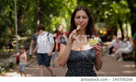 A woman wearing a dark blue polka dot dress stands in a sunny park, holding an ice cream cone and smiling while pedestrians walk by and green trees blur in A woman wearing a dark blue polka dot dress stands in a sunny park, holding an ice cream cone and smiling while pedestrians walk by and green trees blur in 140700266