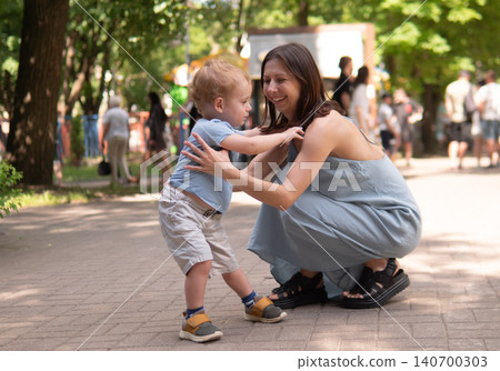 Smiling mother kneels beside her toddler in a sunlit park, reaching toward the child as they interact in a warm, candid moment Smiling mother kneels beside her toddler in a sunlit park, reaching toward the child as they interact in a warm, candid moment 140700303