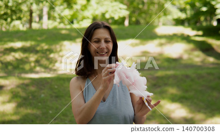 A smiling woman in a sleeveless top stands in a sunlit park, presenting pink fluffy cotton candy on a stick 140700308