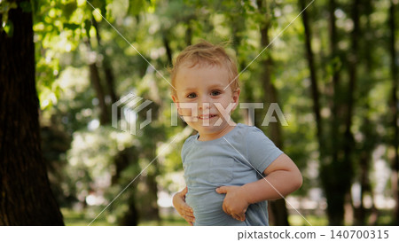 A cheerful young boy stands outdoors in a sunlit park surrounded by trees, wearing a light blue shirt 140700315