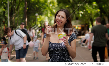 A smiling woman in a polka dot dress enjoys ice cream in a sunny park. People walk in the background as trees and bright afternoon light create a warm, casual A smiling woman in a polka dot dress enjoys ice cream in a sunny park. People walk in the background as trees and bright afternoon light create a warm, casual 140700319