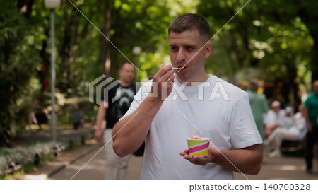 A candid outdoor scene showing a man in a white tshirt enjoying a cup of ice cream while standing along a sunny park path, with trees, pedestrians, and a A candid outdoor scene showing a man in a white tshirt enjoying a cup of ice cream while standing along a sunny park path, with trees, pedestrians, and a 140700328