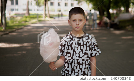 A young boy stands on a sunlit park path, gripping a fluffy pink cotton candy A young boy stands on a sunlit park path, gripping a fluffy pink cotton candy 140700348