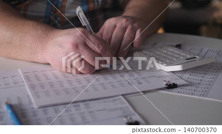 A person is focused on writing down numbers on a sheet of paper while using a calculator. Several papers are spread out on the table, showing different data and notes. A person is focused on writing down numbers on a sheet of paper while using a calculator. Several papers are spread out on the table, showing different data and notes. 140700370