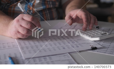 A man writes notes and uses a calculator at a desk filled with documents during the afternoon in an office. The focus is on numbers and calculations for financial tasks. A man writes notes and uses a calculator at a desk filled with documents during the afternoon in an office. The focus is on numbers and calculations for financial tasks. 140700373