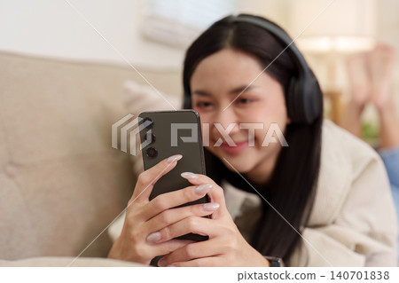 Asian woman with headphones using smartphone on sofa at home Asian woman with headphones using smartphone on sofa at home 140701838