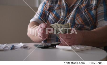 A person sits at a table counting cash. Receipts and a calculator are on the table. The scene captures the act of handling money in a bright room. 140702843