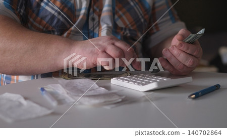 Person uses a calculator to add up bank notes and receipts on a table. This scene takes place indoors, showing a focus on counting and managing money in the afternoon. 140702864