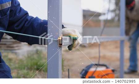 Welder Measuring Alignment On Metal Frame Outdoors, Tape Visible Across Posts, Second Worker Blurred In Background, Gloves Gripping Post, Pragmatic Onsite Workflow, Attention To Straightness Welder Measuring Alignment On Metal Frame Outdoors, Tape Visible Across Posts, Second Worker Blurred In Background, Gloves Gripping Post, Pragmatic Onsite Workflow, Attention To Straightness 140702995