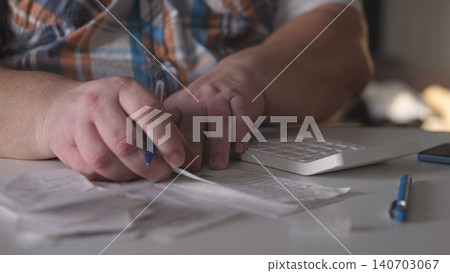 A person sits at a desk with a computer and calculator. They are looking at receipts and writing notes on paper with a pen. The room is bright and organized. 140703067