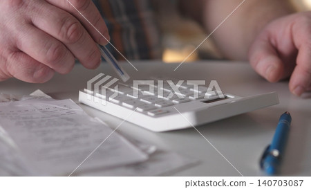 A person presses buttons on a calculator while handling receipts at a desk. Papers are spread out around, and a blue pen rests on the surface nearby as afternoon light shines in. 140703087