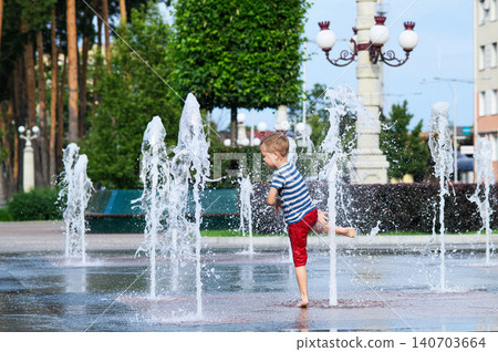 Child playing with water fountain in city square on summer day. Summer lifestyle, joy, freedom and refreshing activity in urban environment. 140703664