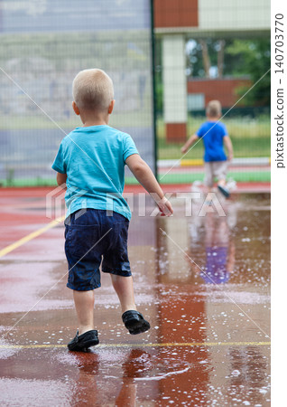 Children riding balance bikes through puddles on wet outdoor sports court after rain. Outdoor lifestyle, childhood activity, freedom and play in urban environment. 140703770
