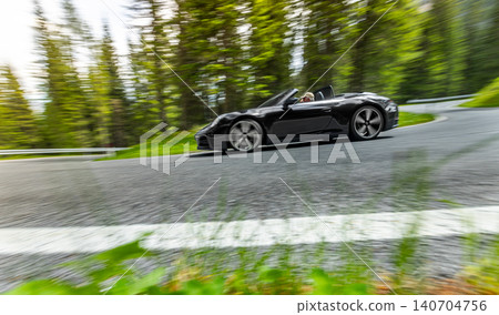 Black convertible sports car driving through a winding mountain road. Motion blur and low angle perspective create a dynamic image of speed, luxury and scenic travel. Black convertible sports car driving through a winding mountain road. Motion blur and low angle perspective create a dynamic image of speed, luxury and scenic travel. 140704756