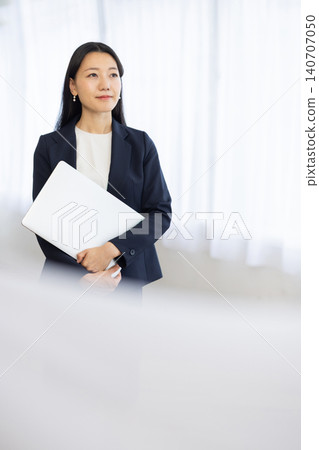 一位年輕的日本女商人面帶微笑地站在窗邊，手裡拿著一台筆記型電腦。 140707050