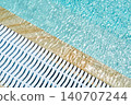 The edge of a pool with blue water and a white plastic drain grate.  140707244